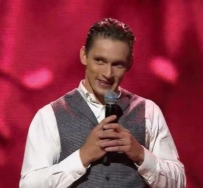 Filip Gurłacz w dynamicznej pozie na scenie telewizyjnej, z tłem sugerującym światła studia i publiczność, oddający energię występu.