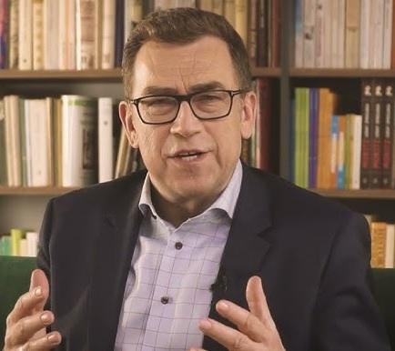 Maciej Orłoś czytający książkę dzieciom, uśmiechnięty, w otoczeniu słuchających go dzieci.