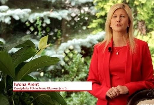 Teresa, mama Iwony Arent, uśmiechnięta przed tradycyjnym domem w skansenie w Olsztynku.
